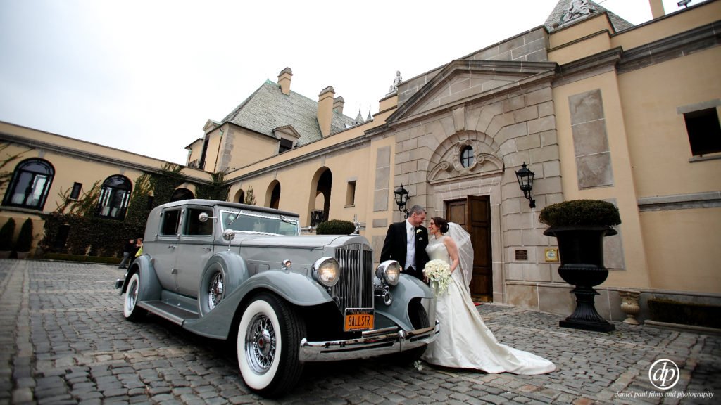 Wedding photo of a bride and groom standing next to a classic car in front of a fancy building.