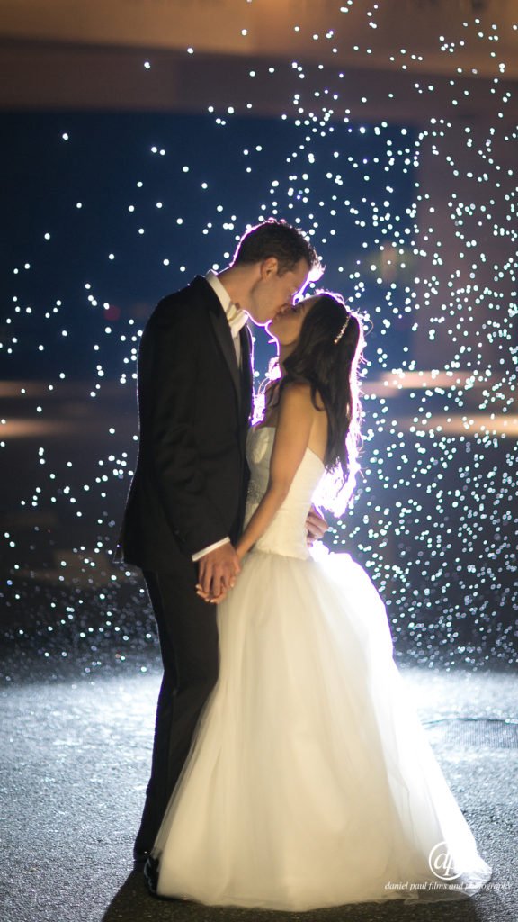 Photo of a bride and groom kissing and holding each other in a spotlight with dramatic lighting effects behind them.