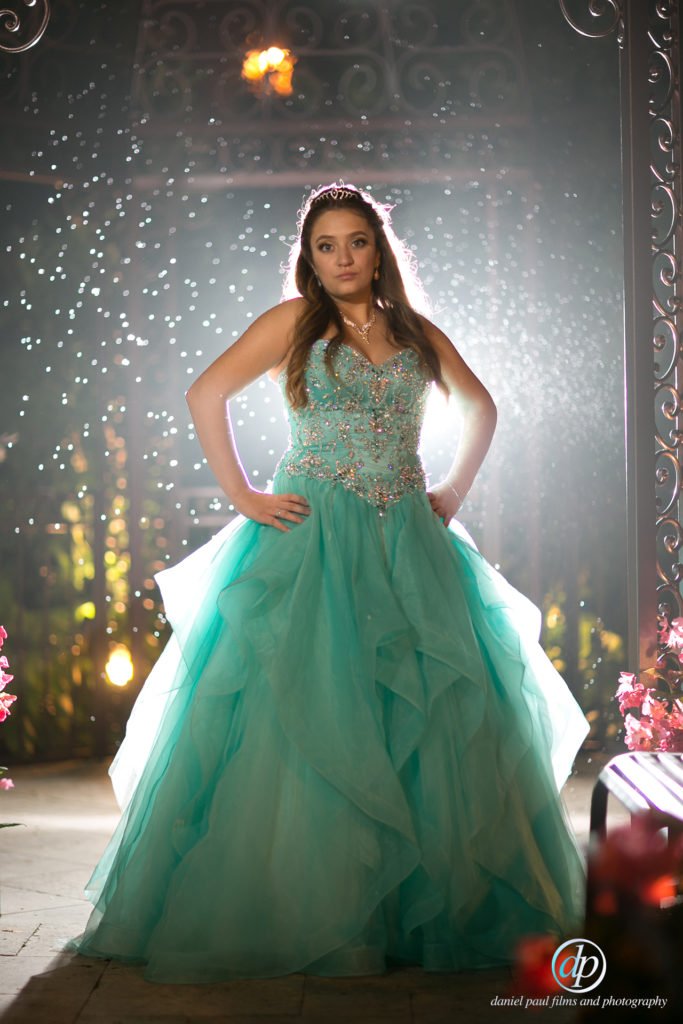 A young 16-year old girl in a sea-foam green dress and tiara in standing in front of a shimmering backdrop.