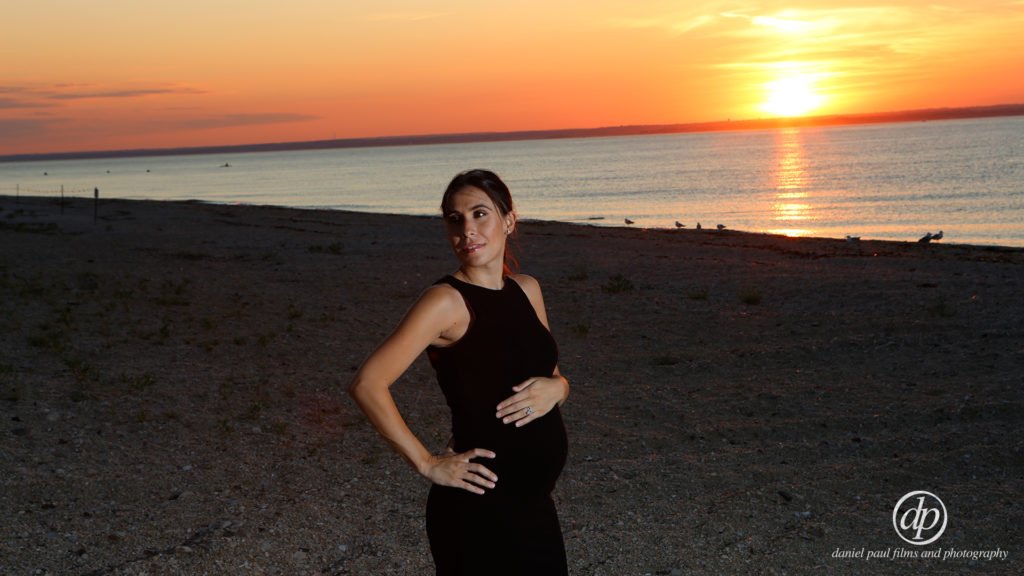 A pregnant woman with her hand cradling her belly at the beach at sunset.